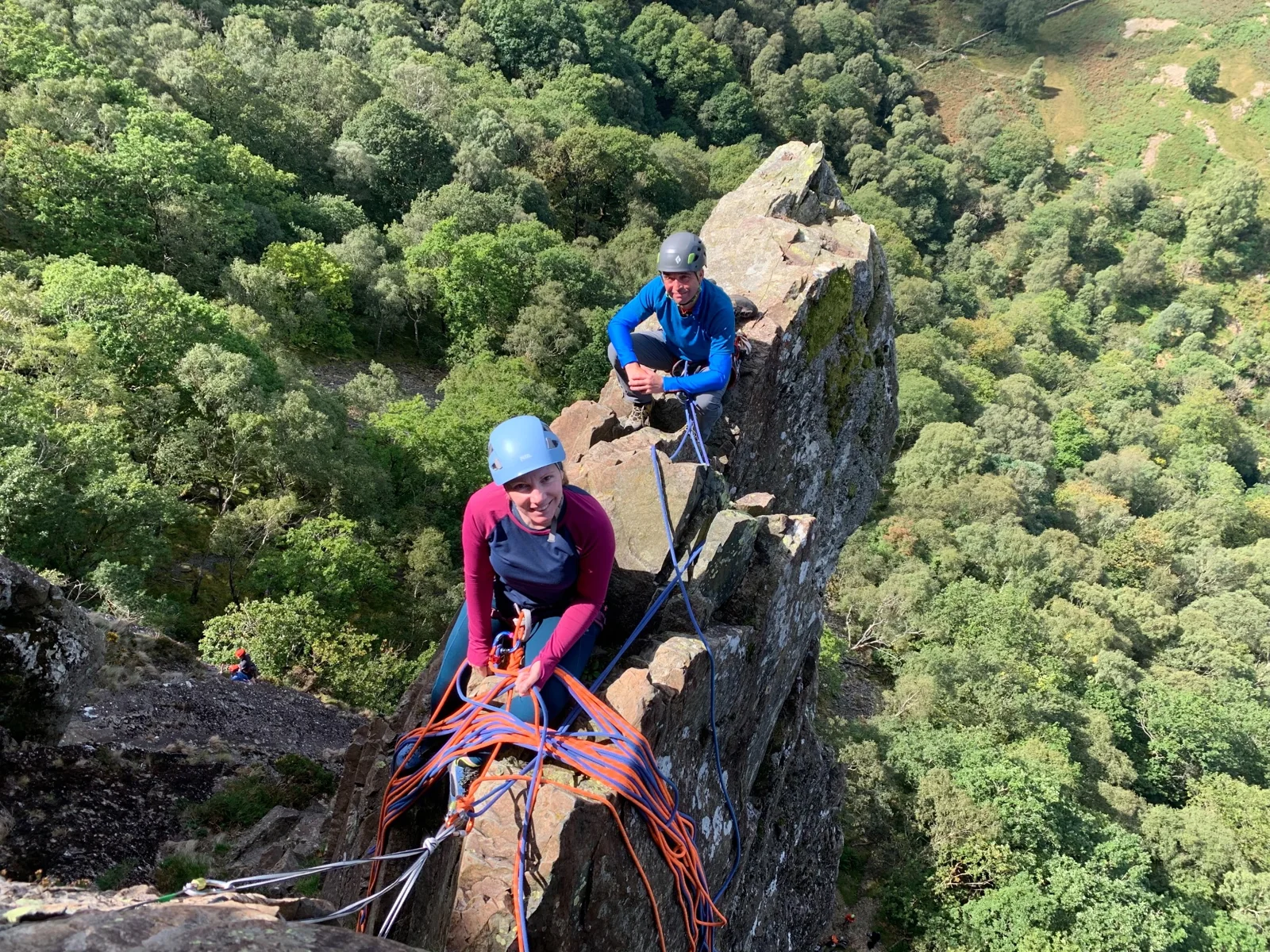 Two helmeted climbers on a narrow rock ridge above woodland