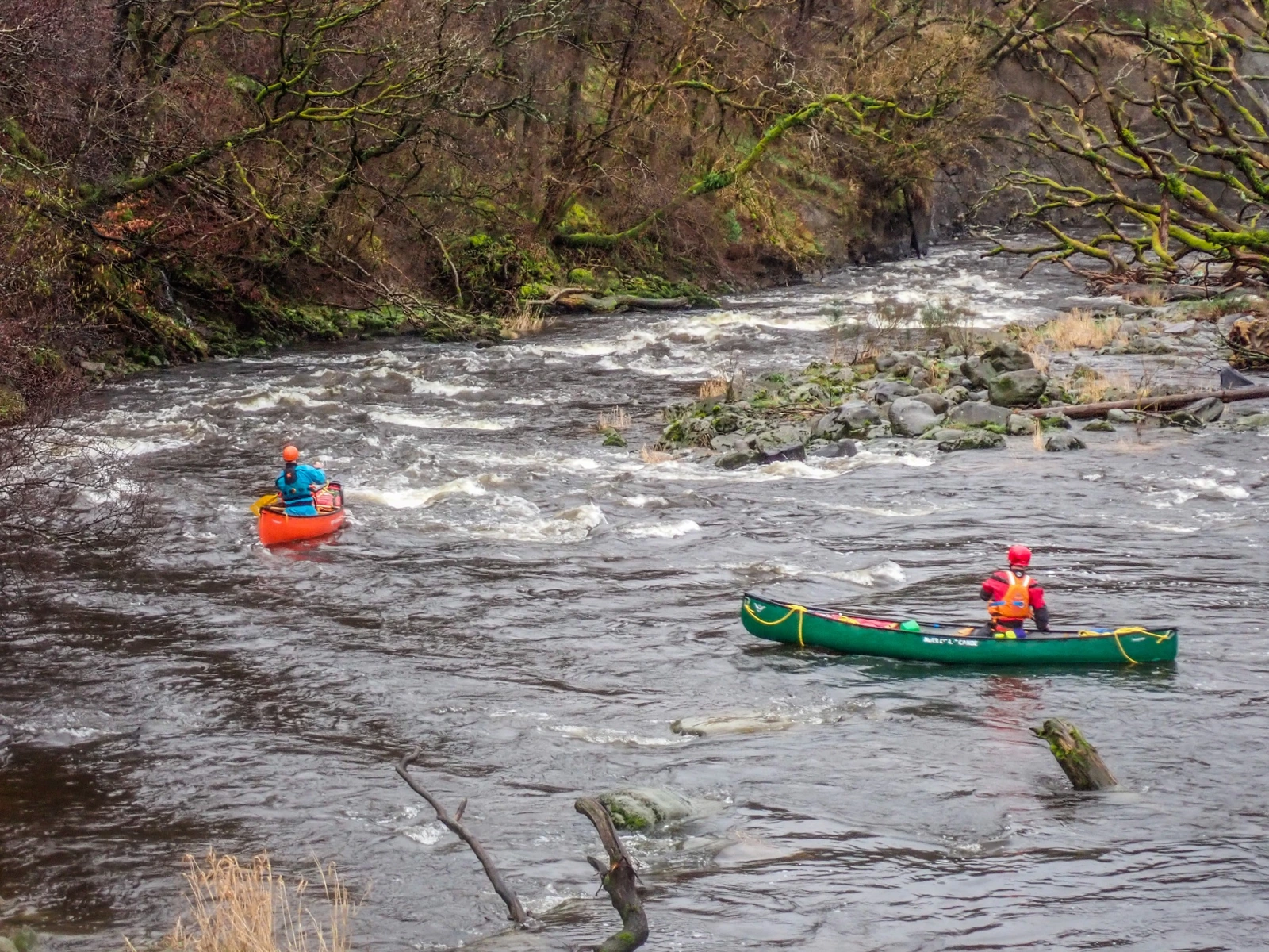Two paddlers in open canoes on a whitewater river through woodland