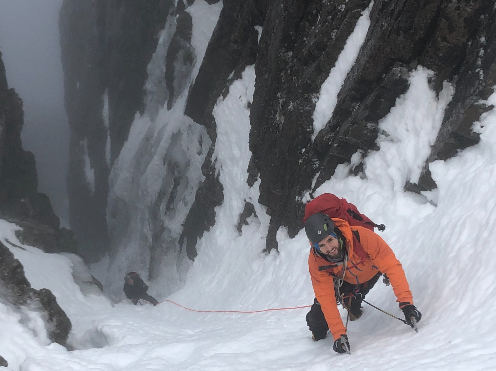 Two winter climbers ascending a snowy gully, with the leading climber in an orange jacket
